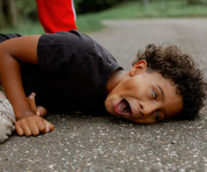 Picture capturing fearful moment when black boy fell on pavement in epileptic fit, lying on side with open mouth and rolling eyes while unseen dad standing behind. Kid with disease, coughing seizure during walk with family.