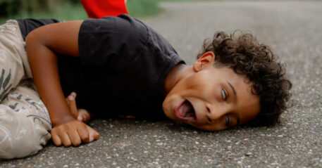 Picture capturing fearful moment when black boy fell on pavement in epileptic fit, lying on side with open mouth and rolling eyes while unseen dad standing behind. Kid with disease, coughing seizure during walk with family.